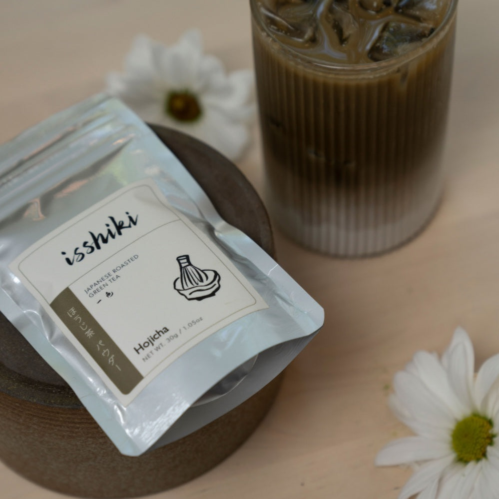 Isshiki Hojicha green tea powder package beside a prepared iced tea drink and white flowers
